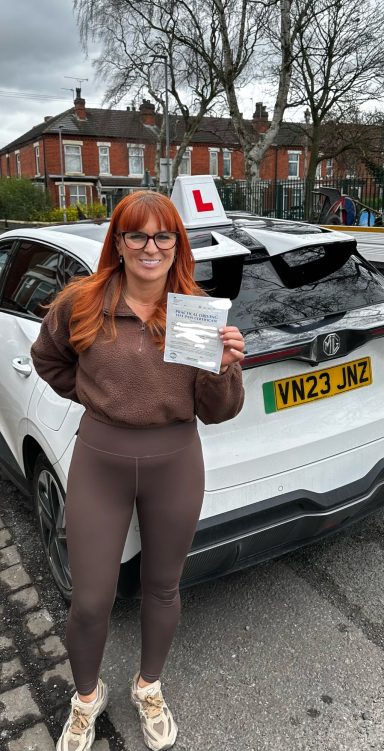 A young woman with long red hair holding a driving test certificate beside a learner car.