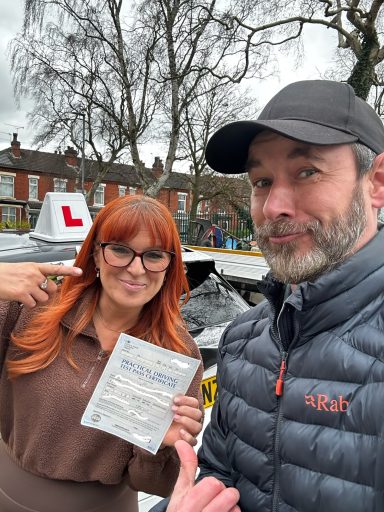 A woman with red hair and glasses poses with a driving test certificate alongside a man.